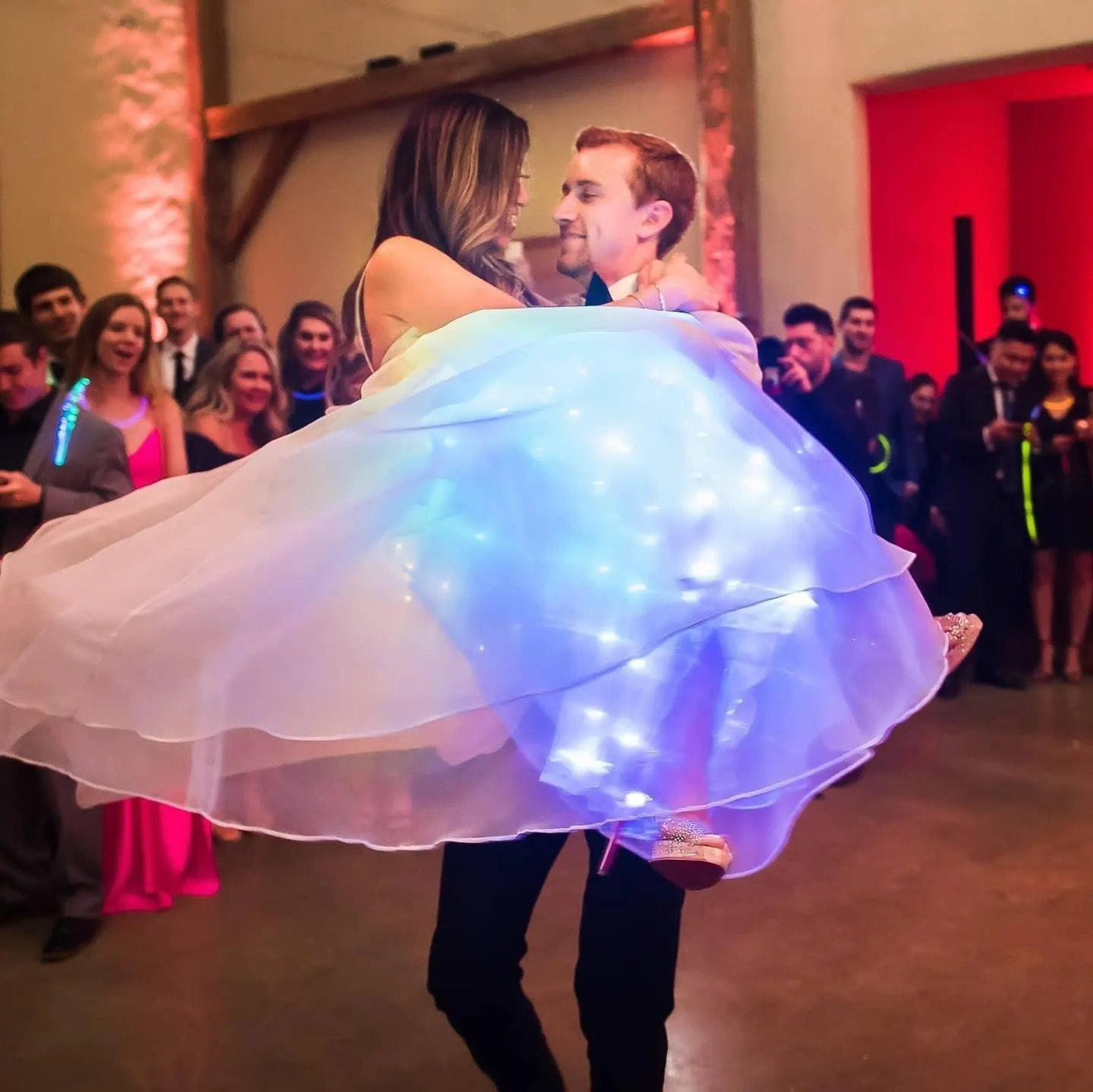 Groom lifting the bride in a lit rainbow gown as guests cheer